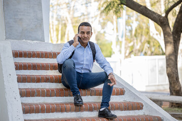 Obraz premium Young latino man sitting on some stairs stairs of a monument on his mobile phone.