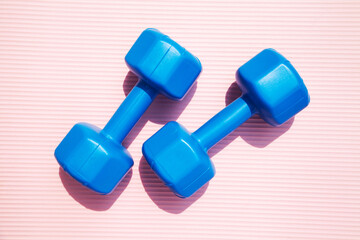 Top view of 2 blue plastic dumbbells on a pink yoga mat. Pair of cheap dumbbells for lifting and workout