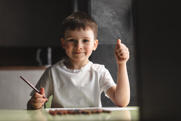 Little preschool boy in white t-shirt draw paint using color pencil and paper sitting at the desk in room at home. Children paint. Kids draw. Creative boy. Boy show thumb up, wow emotions.