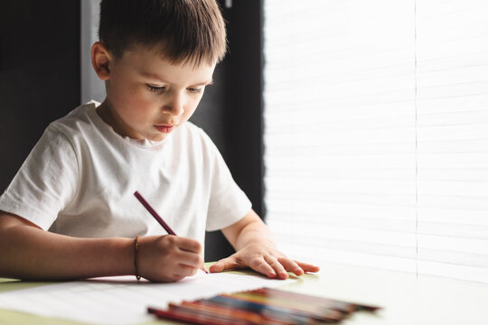 Little preschool boy in white t-shirt draw paint using color pencil and paper sitting at the desk in room at home. Children paint. Kids draw. Creative boy. Childhood, development, education, leisure.