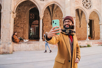 Fototapeta na wymiar Happy young adult man with glasses and beard smiling and taking a selfie portrait on a european city to share on the social media. Caucasian tourist male having fun shooting a photo on a joutney trip