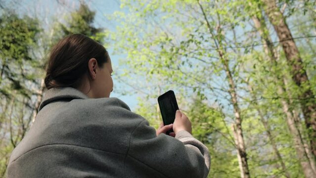 Happy caucasian woman using cell phone taking video and photo in nature with sunlight. Traveller blogger in spring forest. Rest on vacation holiday weekend. Beautiful female enjoying beautiful woods