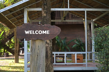 wooden sign "Welcome" in white ink. A brown wooden sign is embroidered on a wooden pole above a large tree near the reception room. Signs are often installed in front of restaurants, shops,homestays.
