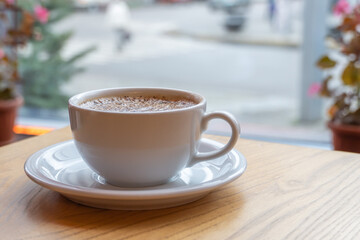 Tasty raf coffee in restaurant on city ​​life background. Still life white cup with hot drink on wooden platform. Coffee steamed and whipped cream with crushed chocolate. Relaxation on terrace in cafe