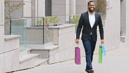 Smiling bearded ethnic male customer client man walking in city with shopping bags buying success buying goods with Black Friday discounts. African American buyer happy with purchases walk outdoors