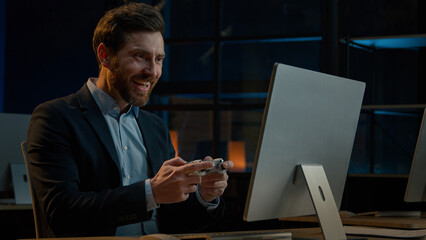 Adult businessman employee working in evening office happy male gamer Caucasian man take break playing console video game on computer with gaming controller using joystick gamepad play virtual match