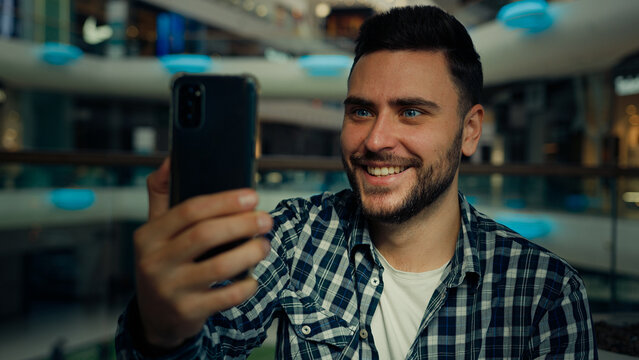 Smiling arabian caucasian male blogger businessman look at mobile phone camera talk video call in shopping mall happy young man chatting online recording blog use modern tech for remote communication