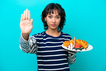 Young Argentinian woman holding waffles isolated on blue background making stop gesture