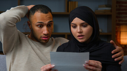 Shocked African Latina man husband with arabian muslim woman wife opening envelope paper letter upset couple multiracial family read bad news correspondence mail spam refusal of bank loan notification