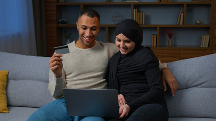 Multiracial couple sit on sofa use laptop purchasing on internet with credit card muslim wife woman in hijab and african american man husband shoppers pay bills online shopping digital payment e-bank