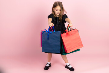 Child shopping. Charming little girl in black stylish summer dress and slight pink makeup, posing with colorful shopping bags. Isolated on pink background.