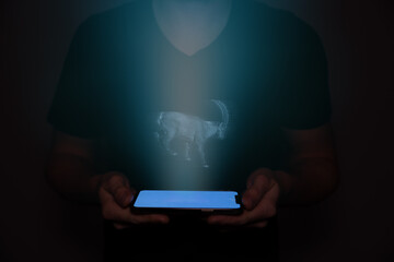 A young man from the future holds a smartphone onto which a capricorn is projected. Future technology.