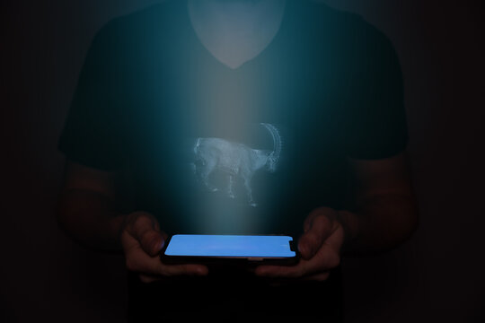 A young man from the future holds a smartphone onto which a capricorn is projected. Future technology.