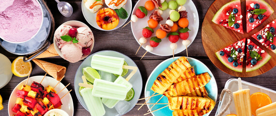 Refreshing summer food table scene. Variety of grilled fruits, ice cream and ice pops. Overhead view on a dark wood banner background.