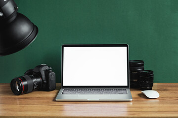 Obraz na płótnie Canvas Modern photographer workplace with computer, DSLR camera and lenses. Laptop with the blank white screen on the wooden office table against dark green wall. Mockup for photo or videomaker advertisement