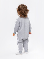 A 2-year-old toddler with curly hair stands on his back in a gray jumpsuit on a white background.