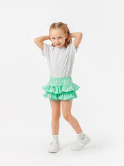A 5-year-old girl in a T-shirt and green skirt smiles with her hands behind her head and bent leg in sneakers on a white background. Looking into the camera