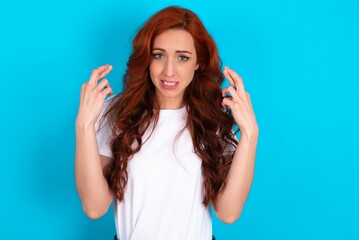 Fototapeta premium young redhead woman wearing white T-shirt over blue background holding fingers crossed with worried expression hoping boss didn't noticed mistakes at work.