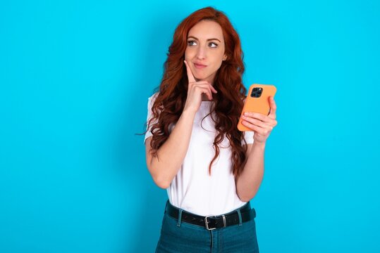 Image of a thinking dreaming young redhead woman wearing white T-shirt over blue background using mobile phone and holding hand on face. Taking decisions and social media concept.