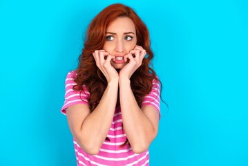 Fototapeta premium Terrified young redhead woman wearing striped T-shirt over blue background looks empty space home alone moonless night