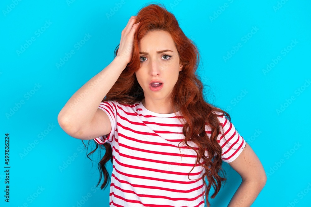 Wall mural Embarrassed young redhead woman wearing striped T-shirt over blue background with shocked expression, expresses great amazement, Puzzled model poses indoor - Wall murals
