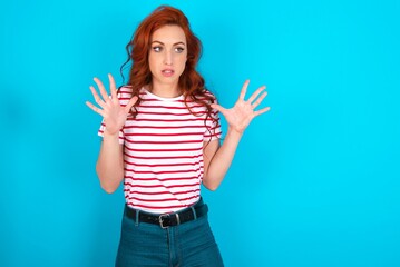 young redhead woman wearing striped T-shirt over blue background shouts loud, keeps eyes opened and hands tense.