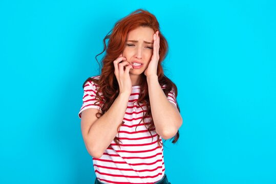 Doleful desperate crying young redhead woman wearing striped T-shirt over blue background , looks stressfully, frowns face, feels lonely and anxious