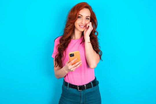 Joyous young redhead woman wearing pink T-shirt poses with mobile phone device, types text message on modern smartphone, watches funny video during free time, enjoys good internet connection,