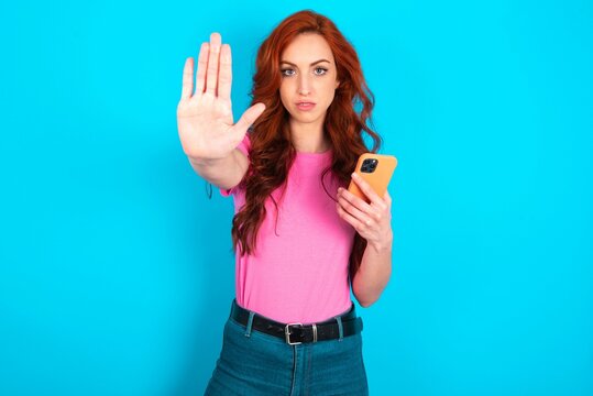 young redhead woman wearing pink T-shirt over blue background using and texting with smartphone with open hand doing stop sign with serious and confident expression, defense gesture