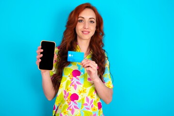 Photo of adorable Young redhead woman wearing colorful shirt over blue background holding credit card and Smartphone. Reserved for online purchases