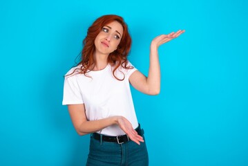 young redhead woman wearing white T-shirt over blue background pointing aside with both hands showing something strange and saying: I don't know what is this. Advertisement concept.