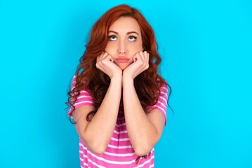 Portrait of sad young redhead woman wearing striped T-shirt over blue background hands face look empty space