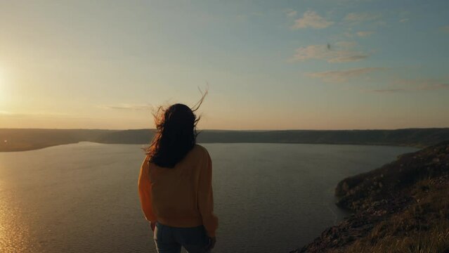 A young woman in a yellow sweater walks to the edge of a cliff above a wide river at sunset, raises her hands in the air, happy and drunk with life, youth and happiness. Watching the sunset 