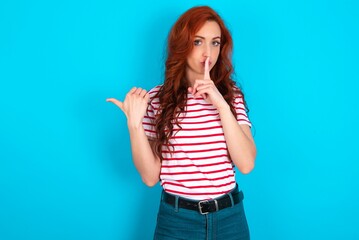 young redhead woman wearing striped T-shirt over blue background asking to be quiet with finger on lips pointing with hand to the side. Silence and secret concept.