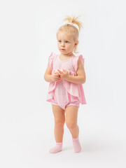 Questioningly serious look. A 1-2-year-old girl in a pink dress and socks stands fingering on a white background.