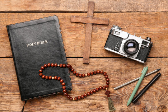 Holy Bible with photo camera, pens, prayer beads and cross on wooden background
