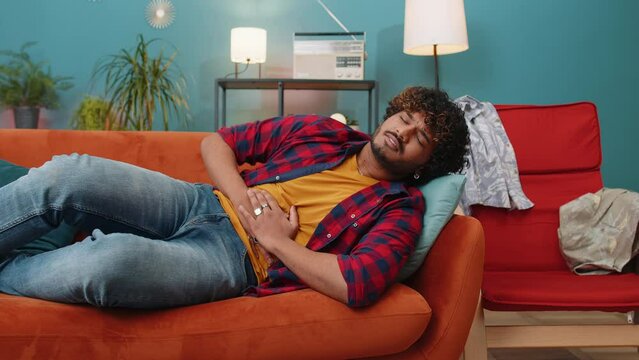 Young indian man lying on sofa feeling sudden strong abdominal stomach ache, gastritis problem. Hindu guy having symptom poisoning diarrhea indigestion peptic ulcer pancreatitis at home apartment room