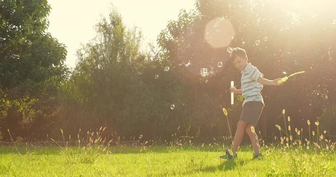Child plays with soap bubbles in nature. Child boy blows soap bubbles, having fun playing on field in rays of sunset. Cute kid smiling, holiday in summer outdoors. Happy childhood concept.