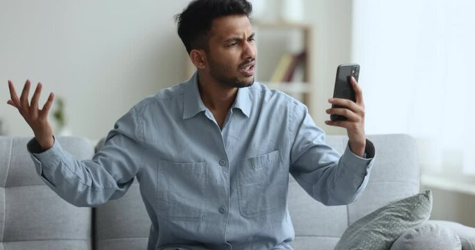 Angry Indian guy sits on couch holds cellphone read unpleasant message, received awful news feels nervous and indignant, experiencing problems with gadget due low battery, no wi-fi Generative AI