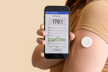 Woman with glucose sensor using mobile phone for measuring blood sugar level on beige background, closeup