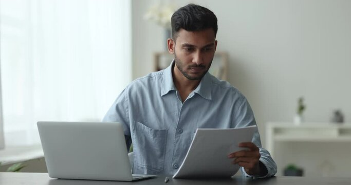 Serious Indian male employee working using laptop, check document terms and conditions, prepare agreement or contract, review details, do paperwork seated at desk. Workflow, legal papers Generative AI