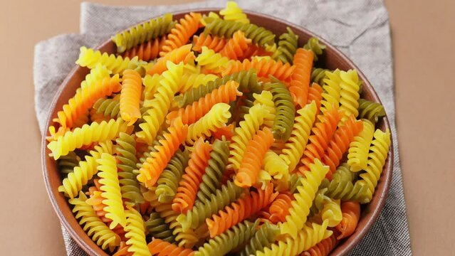 Raw colorful italian fusilli pasta on a beige background. Dry tricolor fusilli pasta on a clay plate. Tilt down camera movement