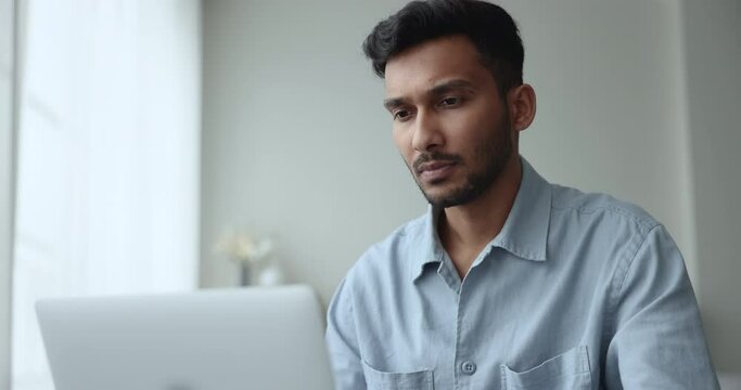 Close up pensive Indian man do remote work on laptop, sit at desk typing on modern device, looks thoughtful, considers decision, response to client, lead chat, correspondence, solve Generative AI