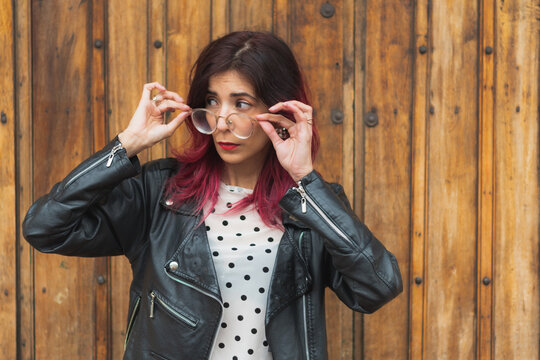 Una mujer  de cabello rojo y chaqueta de cuero negra de pie al frente de una puerta de madera observando hacia la calle con sus manos en las gafas