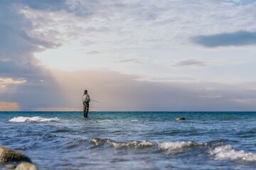 An angler in waterproof trousers is fishing in the Baltic Sea. He is standing on a stone in the water.
