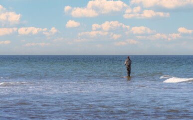 An angler in waterproof trousers is fishing in the Baltic Sea. He is standing on a stone in the water.