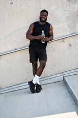 Fit happy sporty young black man standing at urban wall looking at camera feeling confident. Strong African ethnic guy holding water advertising sport street workout training exercises or jogging.