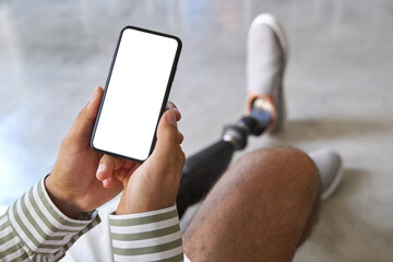 Amputee man with disability leg prosthesis sitting on sofa at home holding cell using mobile phone...