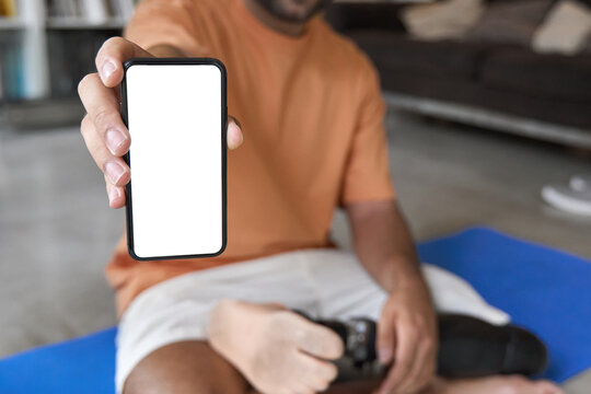 Amputee man with prosthetic leg disability prosthesis sitting on yoga mat at home using phone showing big white mock up screen for advertising digital inclusive sport exercise application.