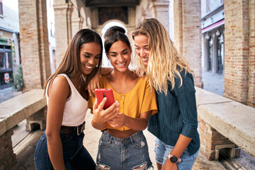 Three happy smiling female friends sharing a tablet computer as they stand close together looking...